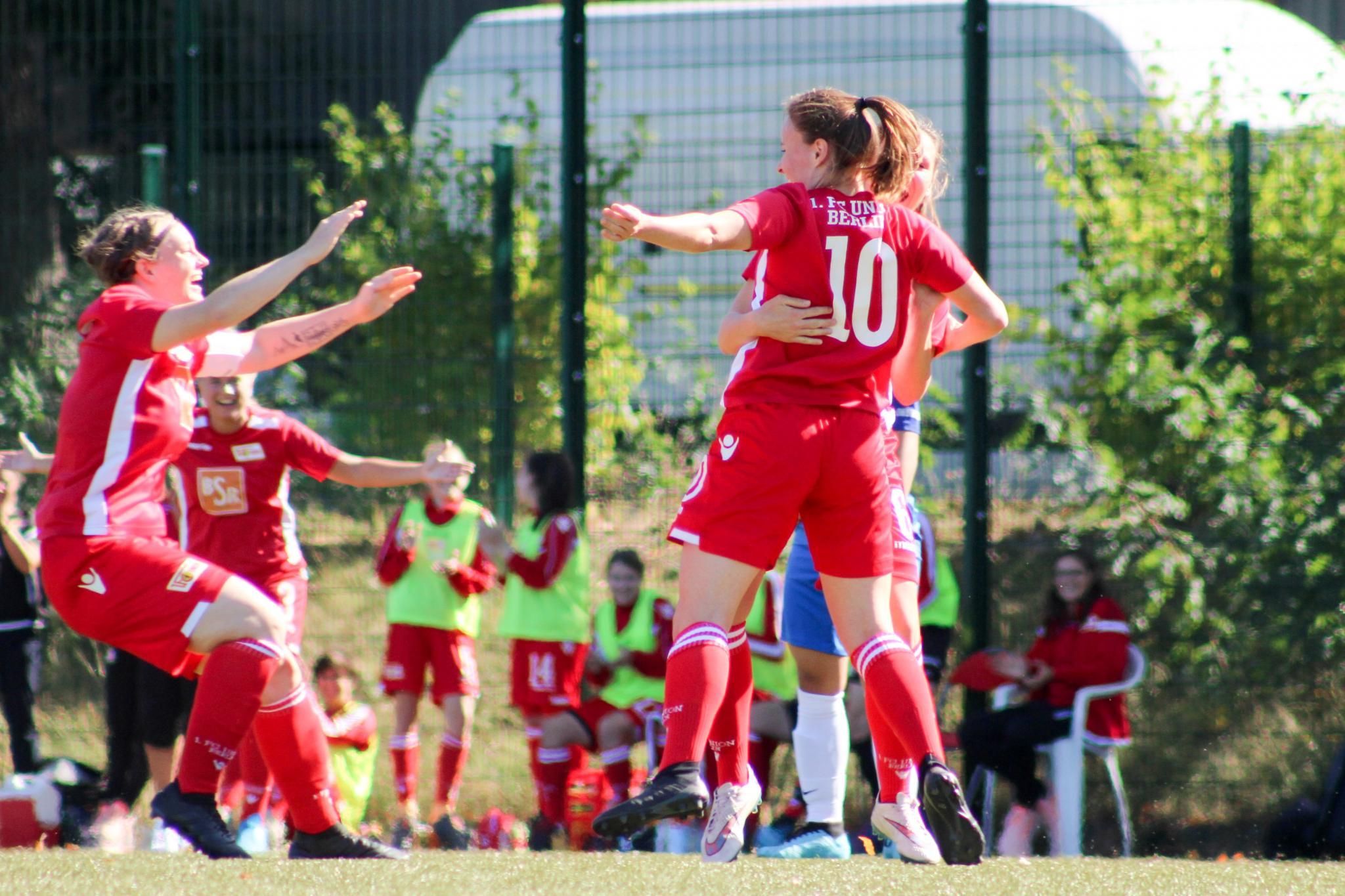 Zwei Fußballspielerinnen in roten Trikots umarmen sich nach einem Tor, während ihre Teamkollegen jubeln.