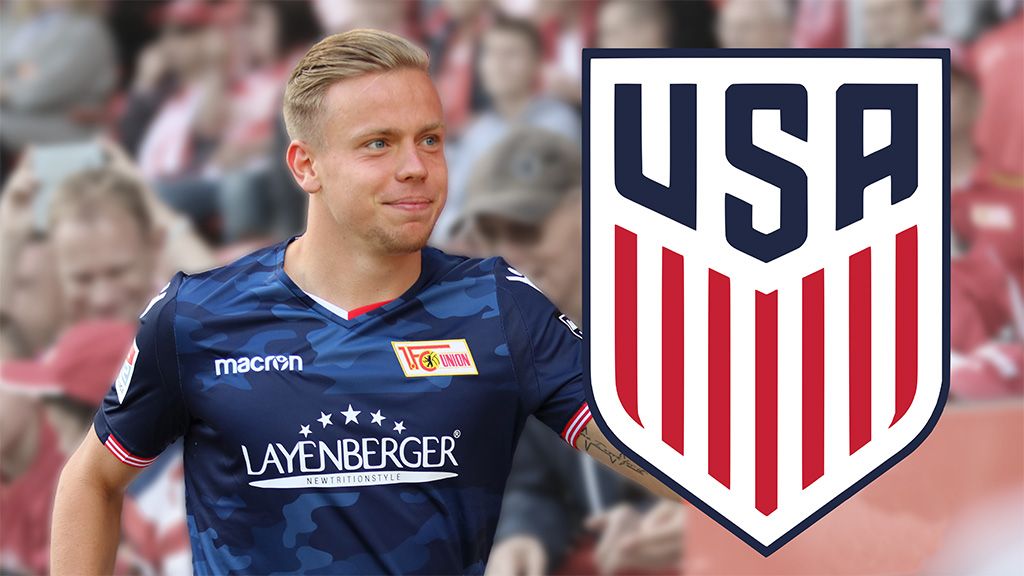 Ein Fußballspieler in einem blauen Trikot steht neben dem USA-Logo. Hintergrund mit Fans im Stadion.