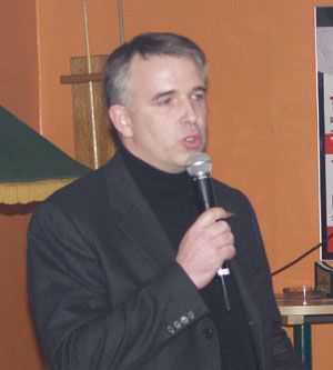 Mann in schwarzem Anzug und Rollkragenpullover spricht mit Mikrofon in der Hand vor orangefarbener Wand.