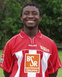 Lächelnder junger Mann in einem roten Fußballtrikot mit Logo, stehend vor grünem Hintergrund.