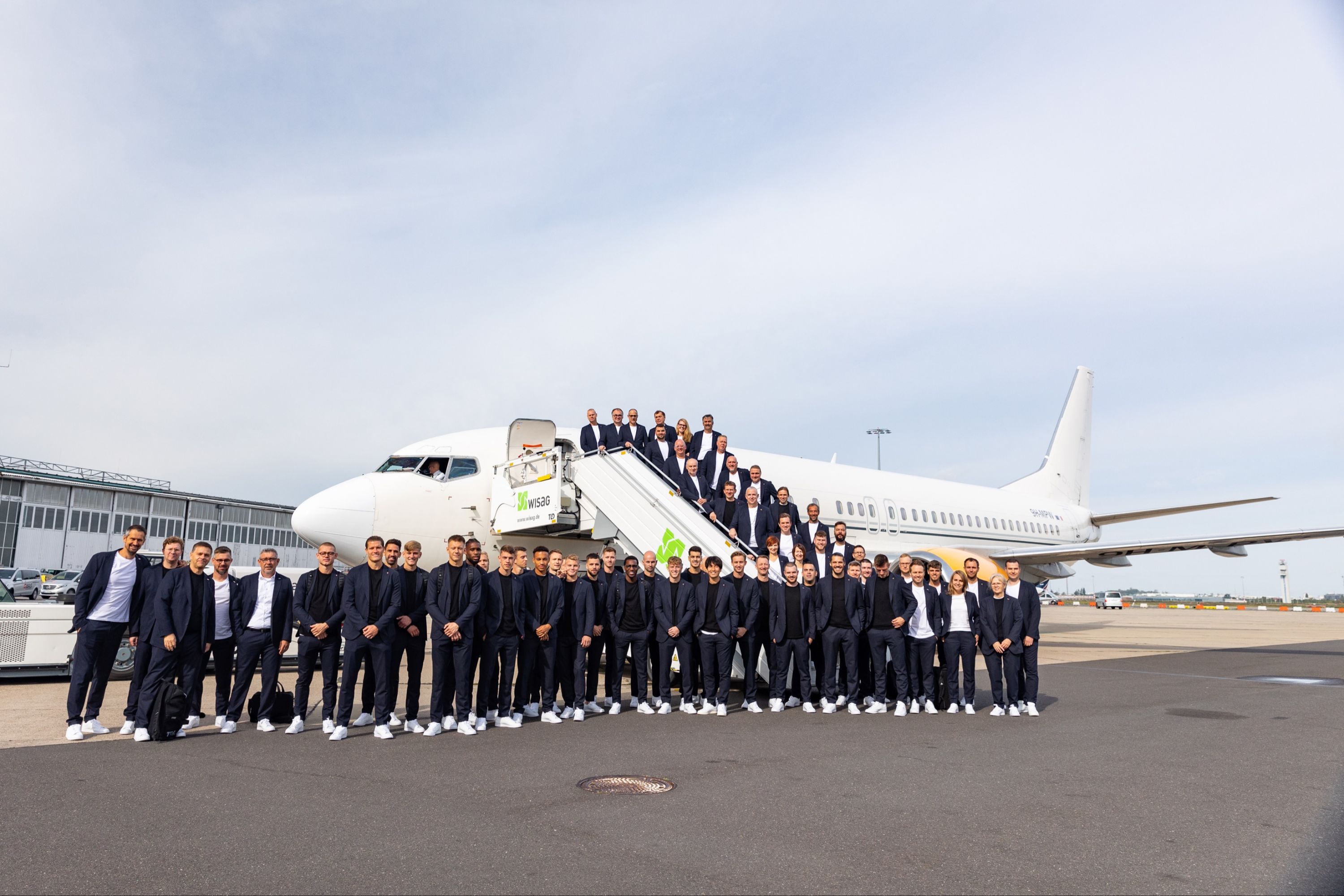 A large group of people in identical black clothing stands in front of an airplane and smiles at the camera.