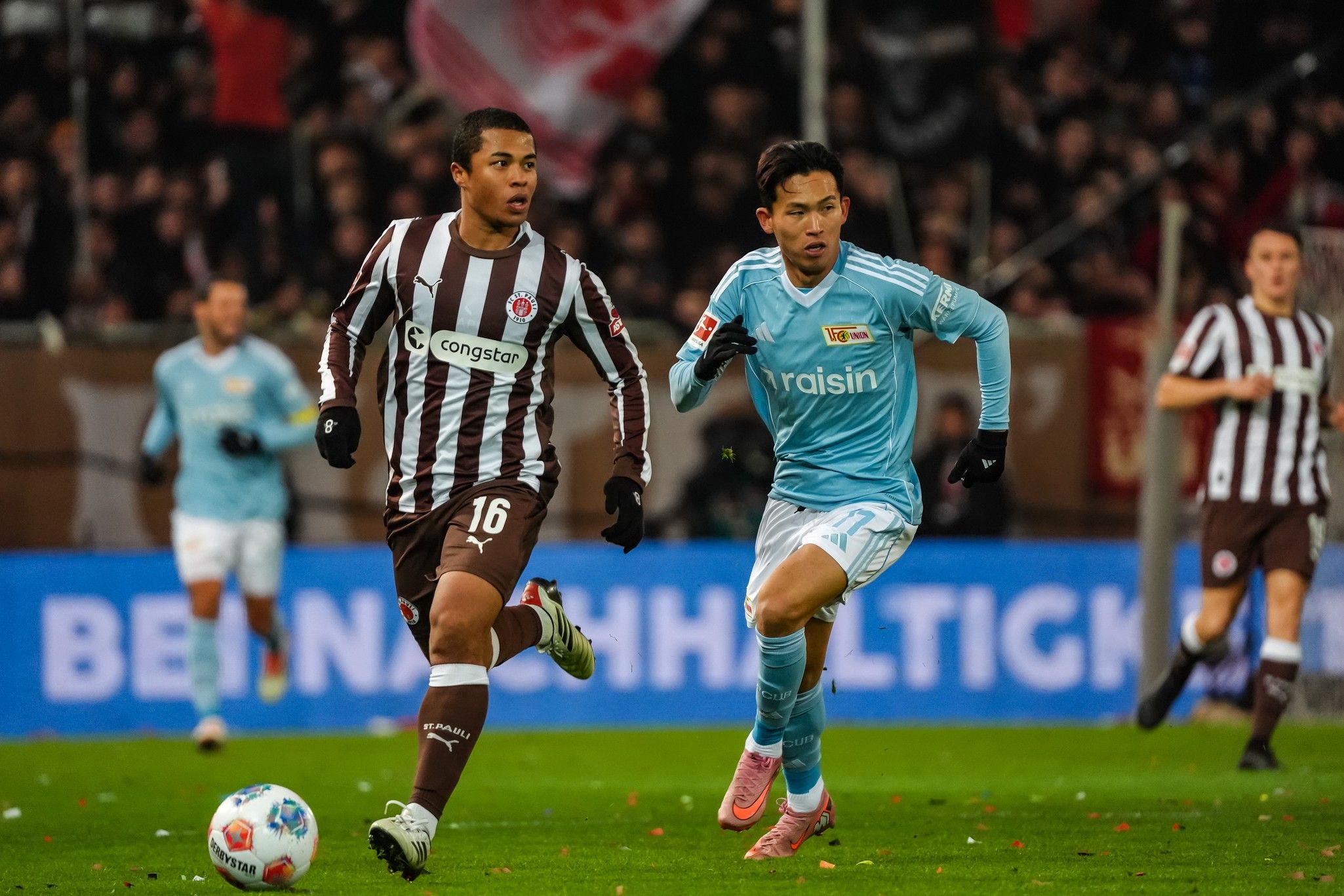 Zwei Fußballspieler in Aktion während eines Spiels, der Spieler mit der Nummer 16 in gestreiftem Trikot und ein anderer in himmelblauem Trikot.