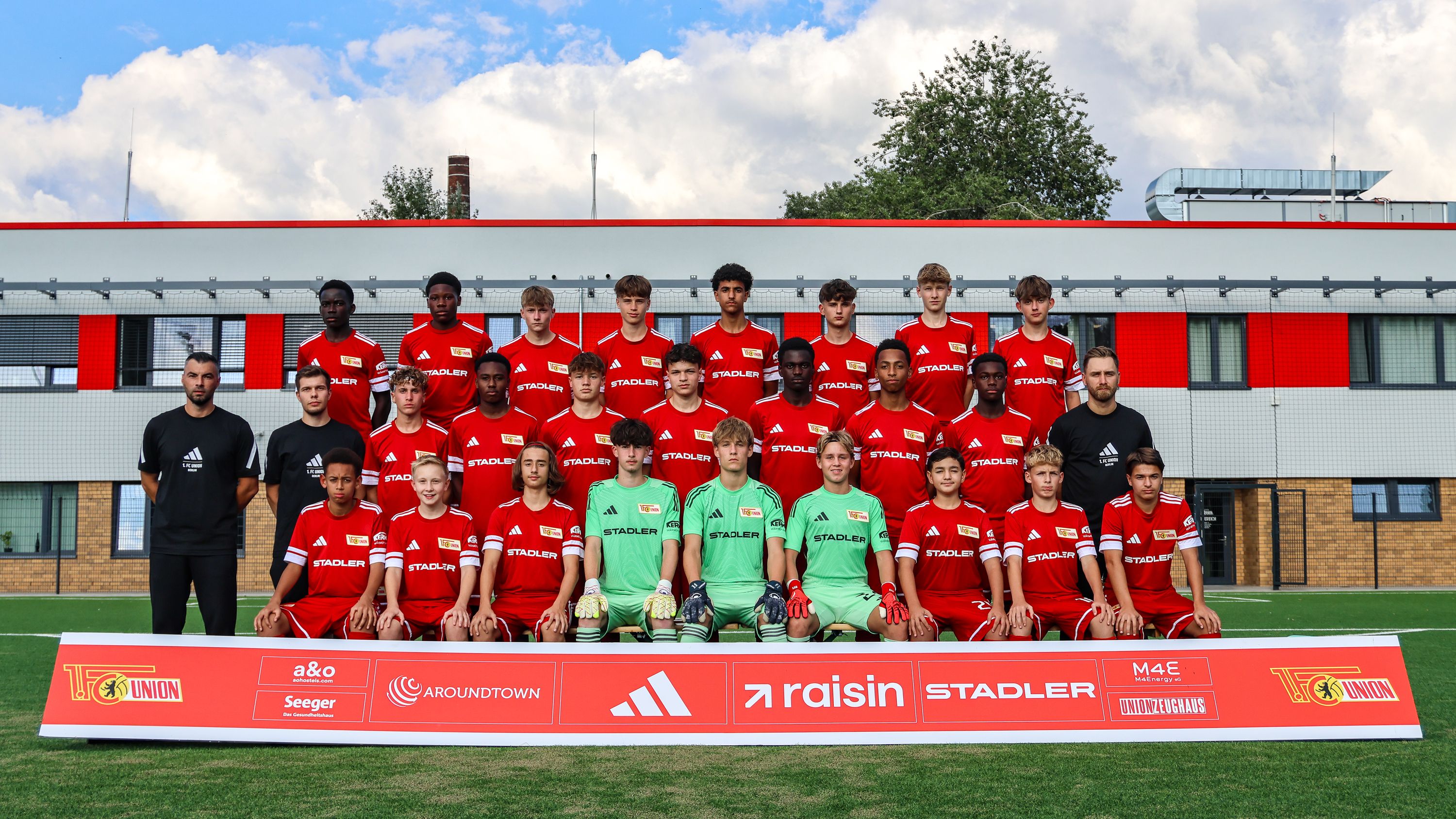 Mannschaftsfoto einer Jugendfußballmannschaft in roten Trikots, stehend und kniend auf einem Grasfeld vor einem Sportgebäude.