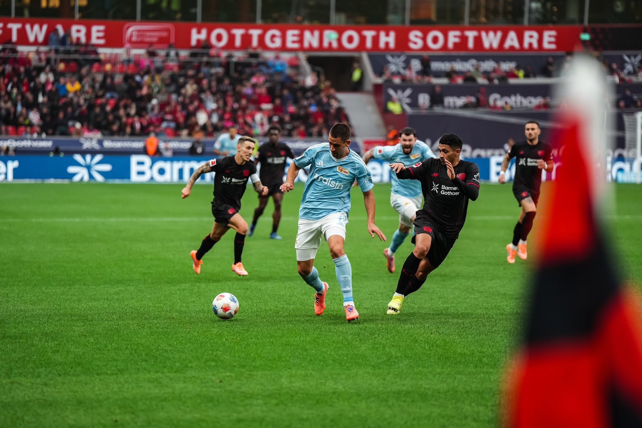 Ein Spieler in blau chase einen Ball, während mehrere gegnerische Spieler in schwarz ihn verfolgen. Stadionatmosphäre im Hintergrund.