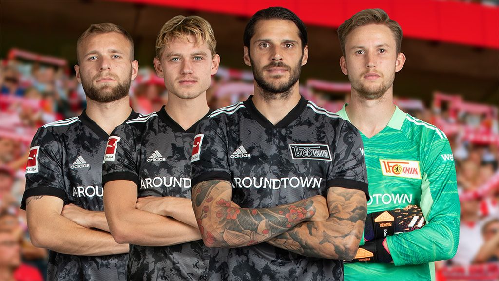 Vier Fußballspieler in schwarzen Trikots stehen zusammen mit armschlüssiger Pose vor einem Stadionhintergrund.