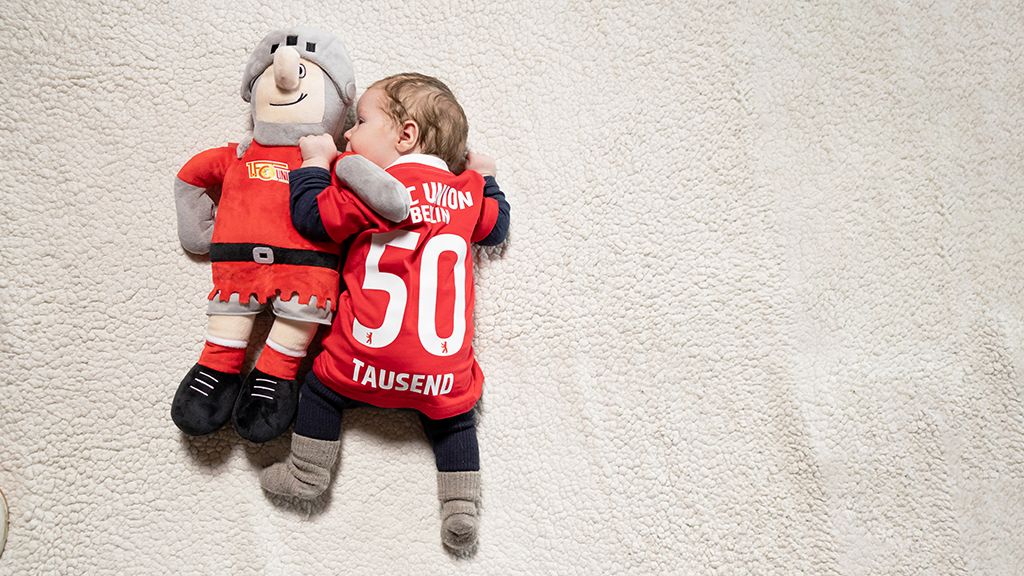 Ein Baby in einem roten Trikot liegt neben einem Stofftier in ähnlicher Farben. Beide liegen auf einem weichen, hellen Untergrund.