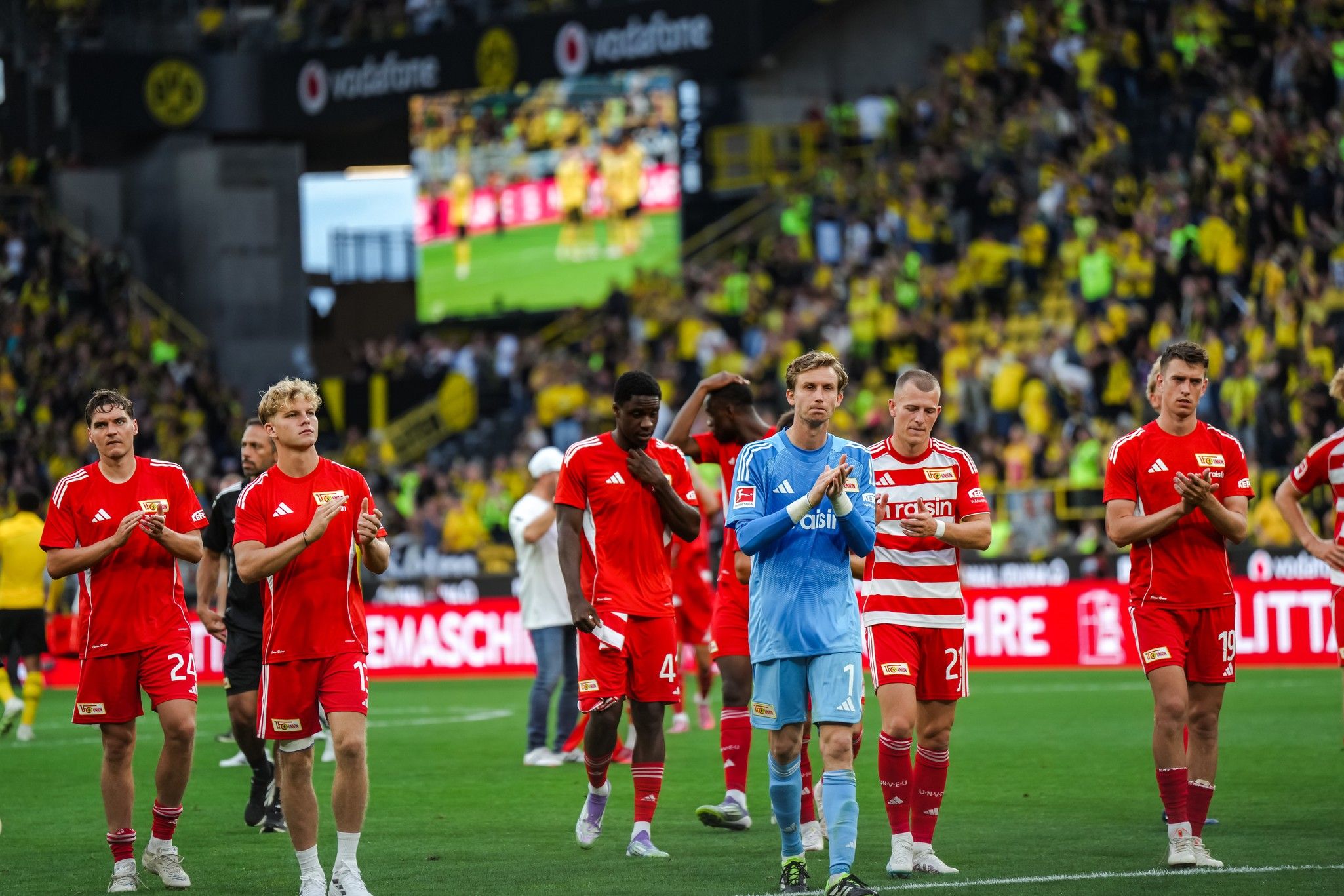 Spieler des 1. FC Union Berlin applaudieren nach einem Spiel, im Hintergrund eine jubelnde Menge und eine Videoleinwand.