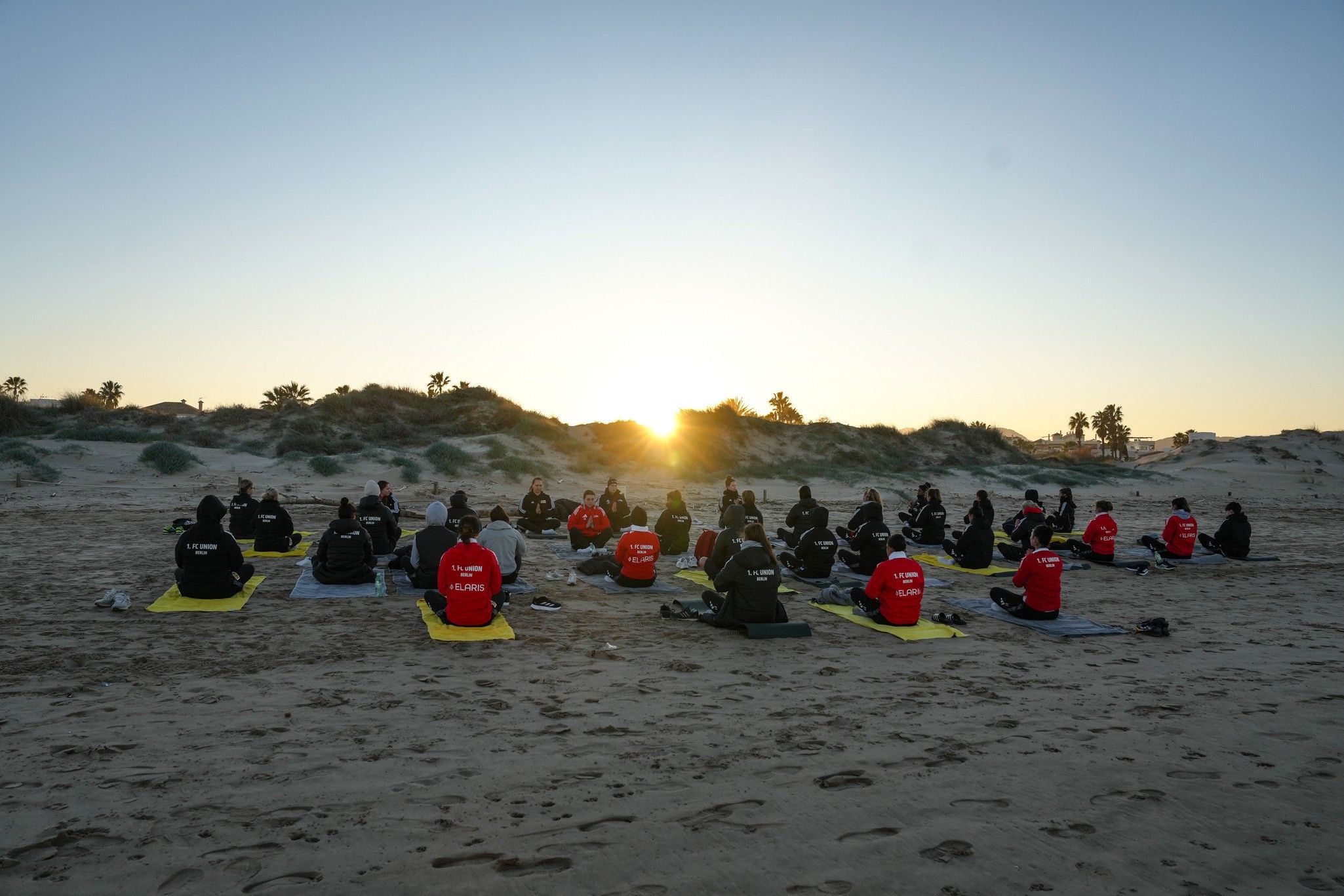 Gruppensitzung auf einem Strand bei Sonnenaufgang, Teilnehmer in schwarzen und roten Sweatshirts, auf Yoga-Matten sitzend.