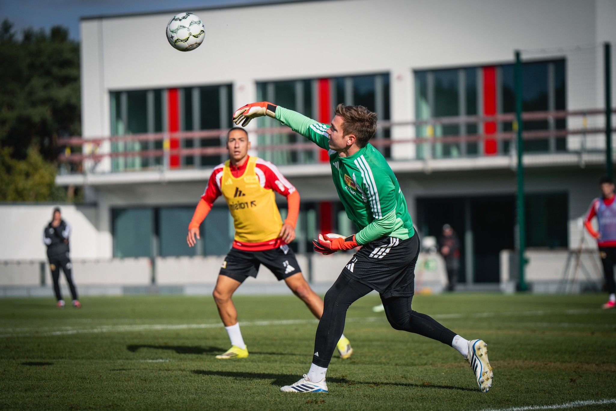 Torwart in grünem Trikot fängt einen Ball während des Trainings, ein Spieler im Hintergrund übt Anspiel.