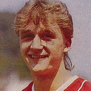 Ein junger Mann mit lockigem, blondem Haar lächelt in die Kamera, trägt ein rotes Sporttrikot.