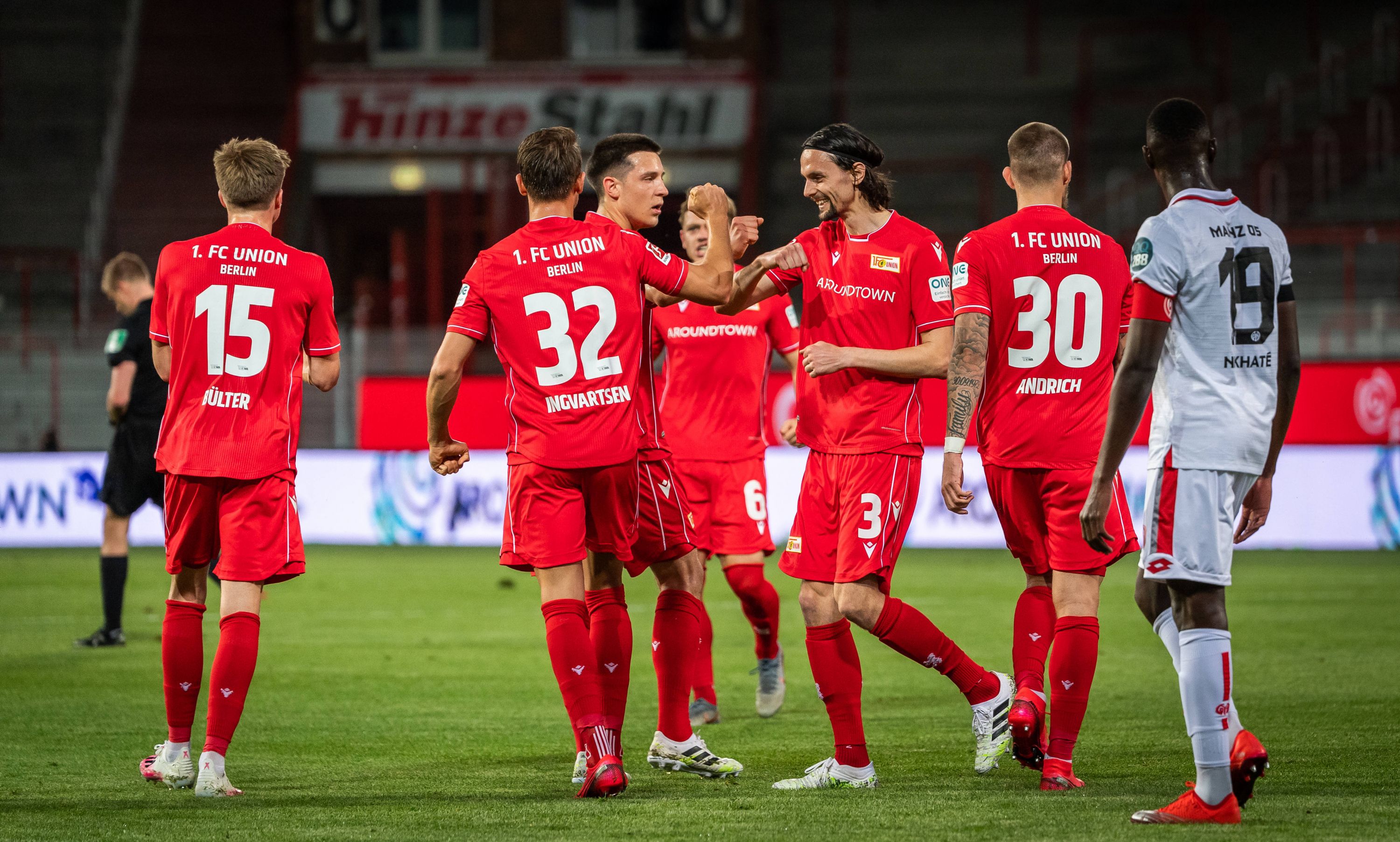 Fußballmannschaft in roten Trikots feiert einen Treffer auf dem Feld. Spieler stehen zusammen, einige mit erhobenen Händen.