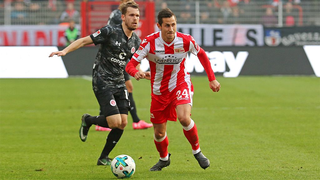 A football player in Union Berlin's kit dribbles the ball while an opposing player chases him.