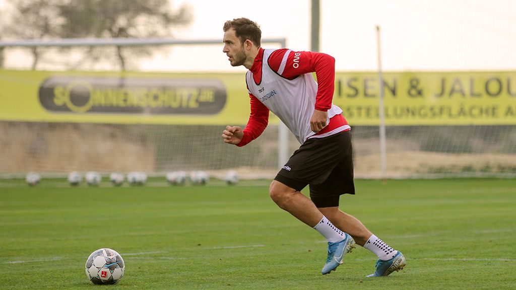 Ein Fußballspieler in Trainingskleidung läuft mit einem Ball auf einem grünen Rasenfeld, im Hintergrund ein präpariertes Tor.