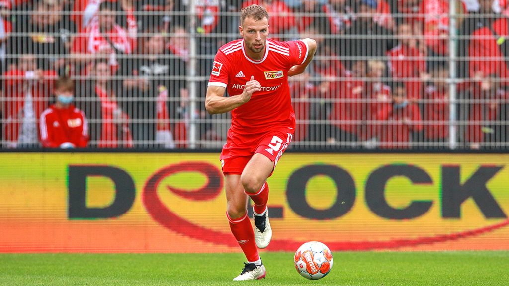 A soccer player in a red jersey runs with the ball on a field, surrounded by cheering fans.