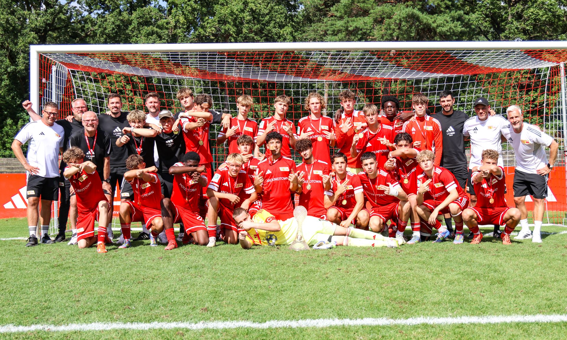 Eine Gruppe von Fußballspielern in roten Trikots posiert gemeinsam auf einem Platz vor einem Tor, einige zeigen den Siegzeichen.