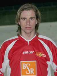 Ein Fußballspieler trägt ein rotes Trikot mit dem Logo seines Vereins und posiert für die Kamera.