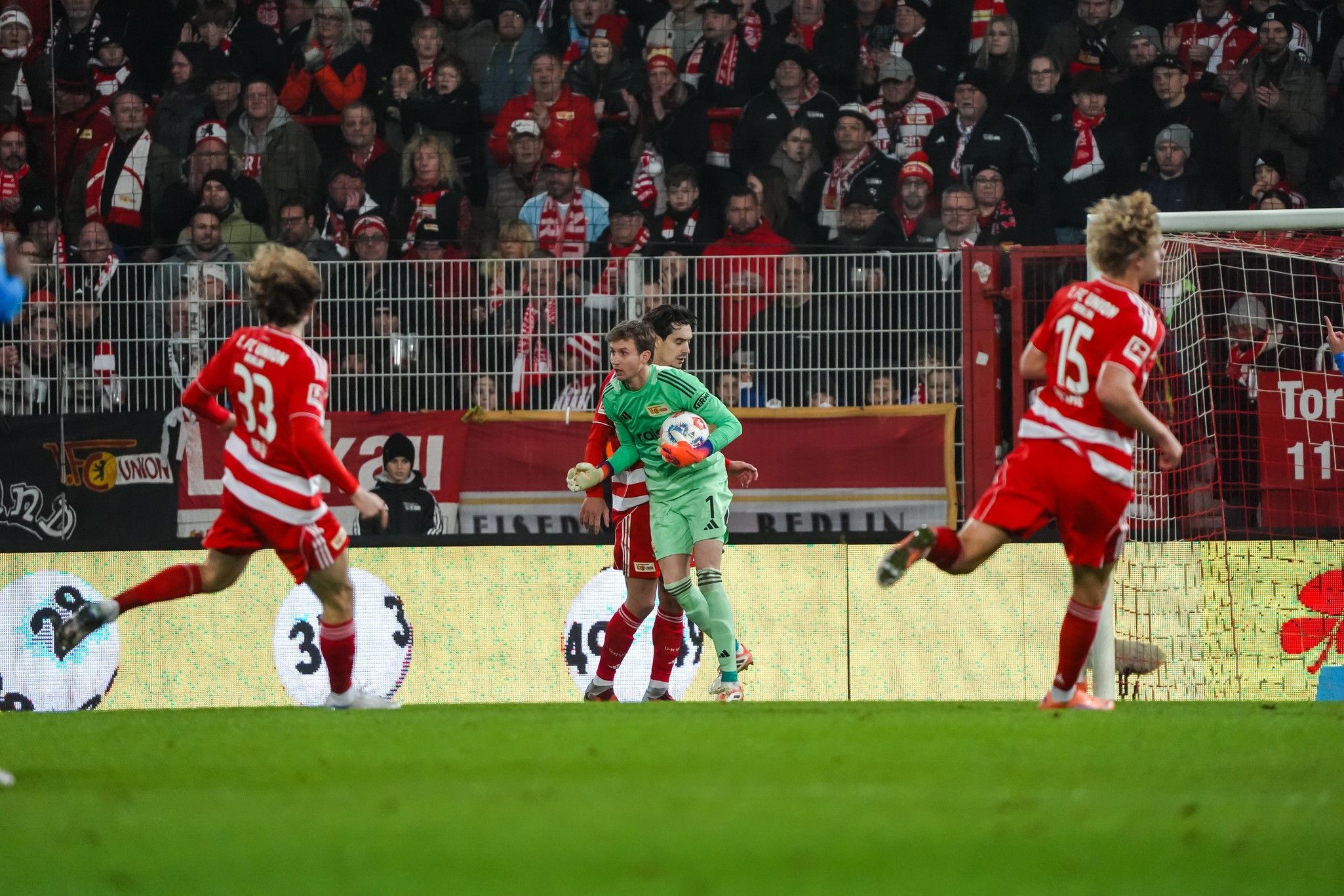 Torwart eines Fußballteams hält den Ball, während Spieler in rot und weiß im Hintergrund jubeln. Fans in der Tribüne sind sichtbar.