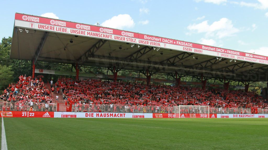 Stadionatmosphäre in einem Fußballspiel mit einer vollen Tribüne in roter Kleidung und einer klaren blauen Himmel.