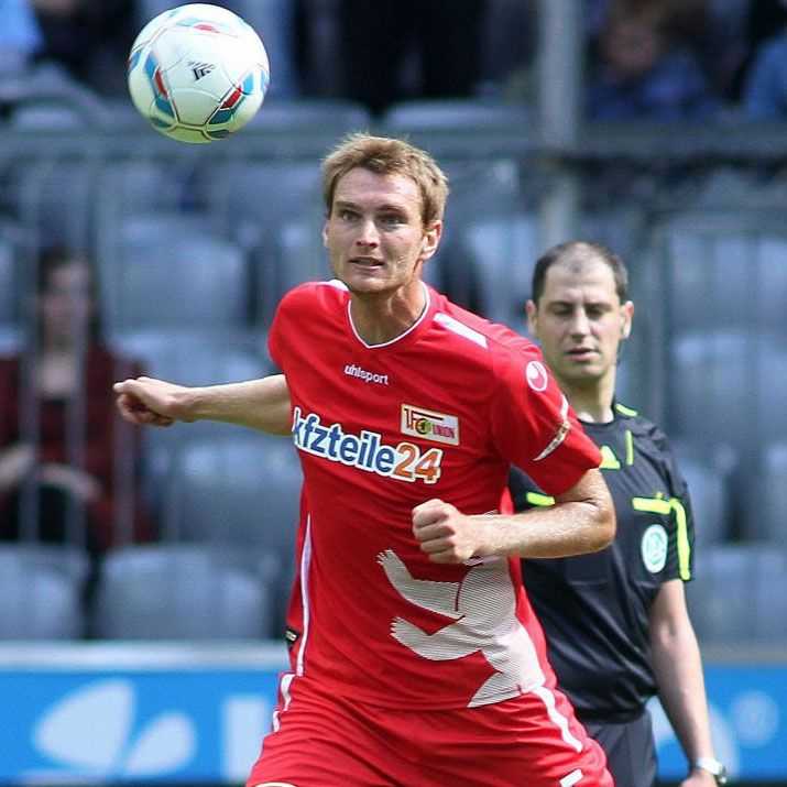 Ein Fußballspieler in roter Kleidung mit einer Fußballicon, der einen Ball in der Luft verfolgt, während ein Schiedsrichter im Hintergrund steht.