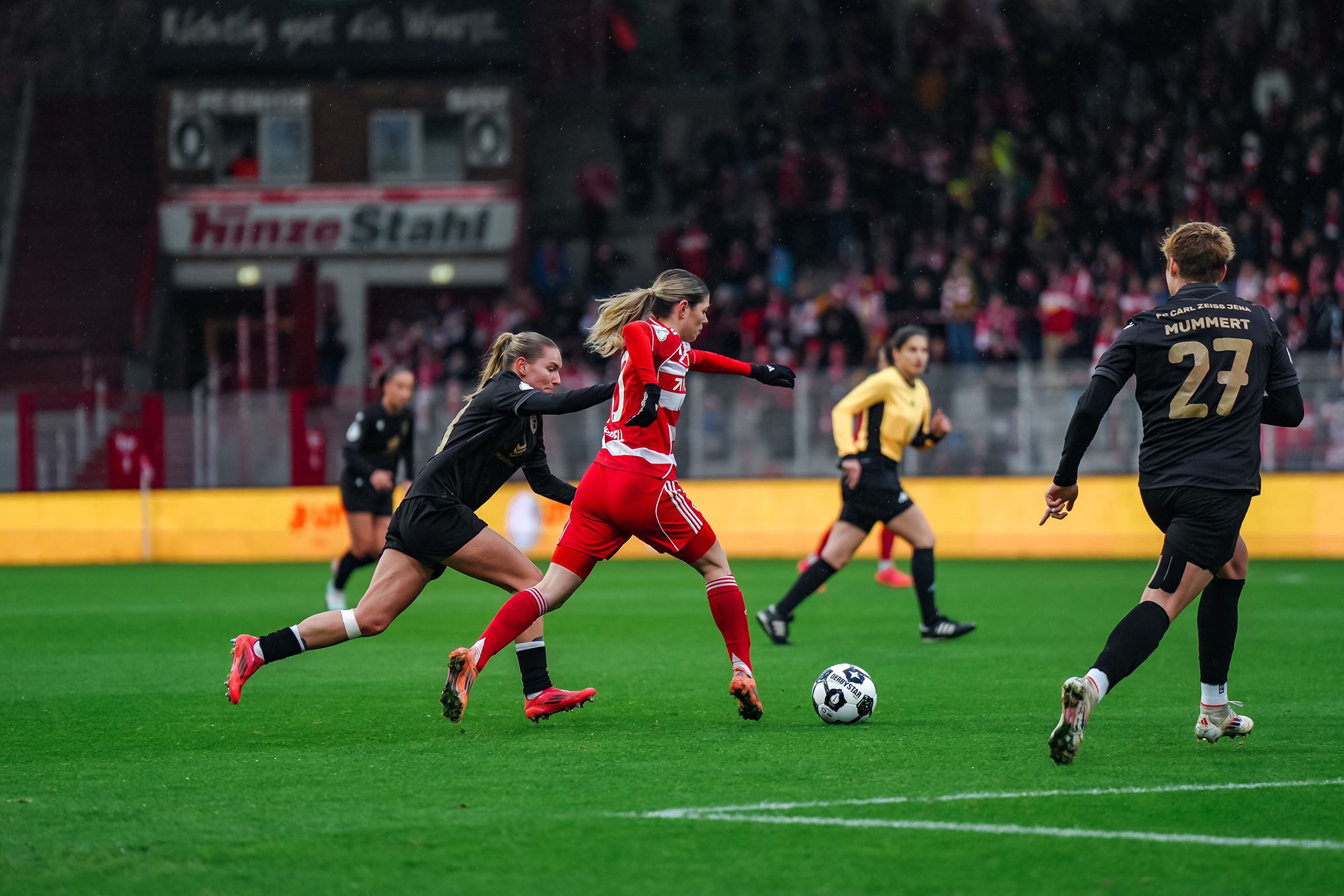 Eine Fußballspielerin in rotem Trikot dribbelt mit dem Ball, während eine Gegnerin in schwarz versucht, sie zu stoppen.