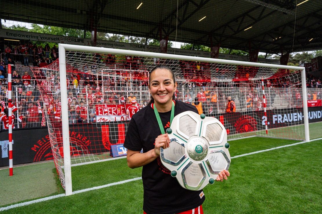 Eine lächelnde Frau hält eine Meisterschale vor einem Fußballtor in einem Stadion, umgeben von Fans und roten Fanfaren.