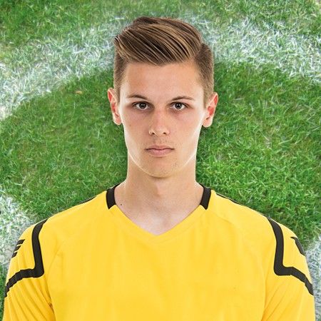 Junger Fußballspieler im gelben Trikot mit neutralem Ausdruck vor einem grünen Fußballplatzhintergrund.