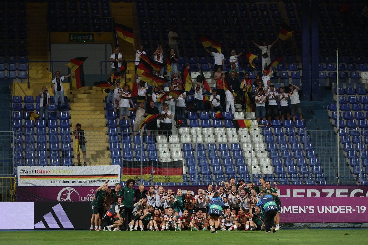 Feiernde deutsche Fußballmannschaft mit Fans in einem Stadion, im Hintergrund sind Deutschland-Fahnen und ein großes "A" sichtbar.
