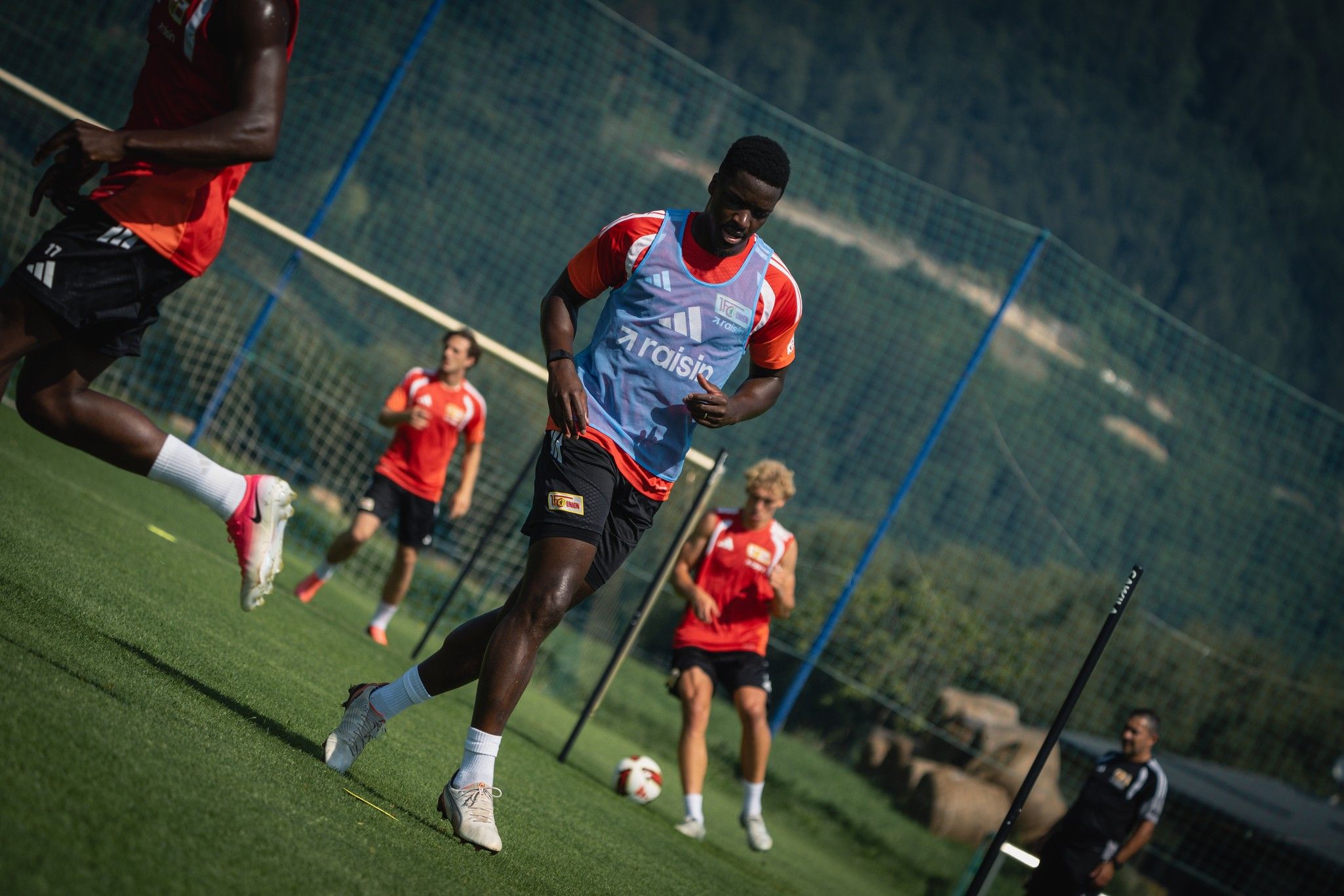 Fußballspieler in Trainingskleidung auf dem Platz, umgeben von Teamkollegen im Hintergrund.