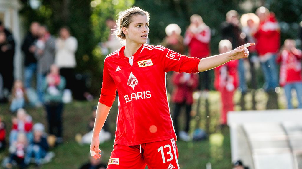 Eine Fußballspielerin in rotem Trikot mit der Nummer 13 zeigt auf das Spielfeld, während Zuschauer im Hintergrund zu sehen sind.
