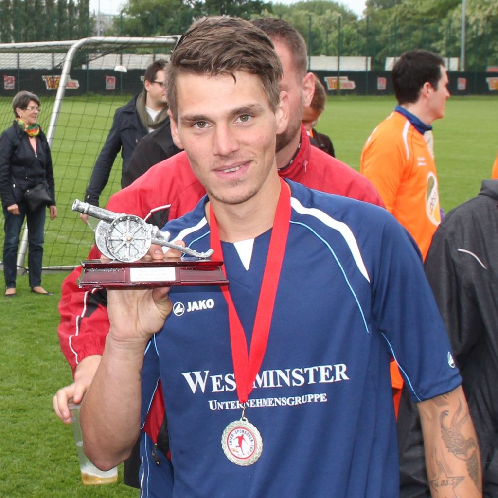 Ein junger Mann in einem blauen Sporttrikot hält stolz einen pokalähnlichen Preis in der Hand. Hintergrund mit Fußballfeld und Spielern.