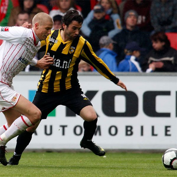 Zwei Fußballspieler im Wettkampf um den Ball, einer in einer weißen und der andere in einer schwarz-gelben Uniform. Zuschauer im Hintergrund.