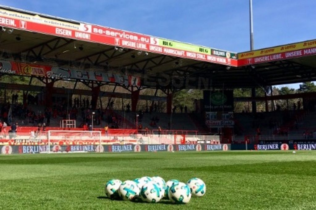 Nahaufnahme von einem Fußballfeld mit mehreren Fußbällen auf dem Rasen, im Hintergrund das Stadion und Zuschauertribünen.