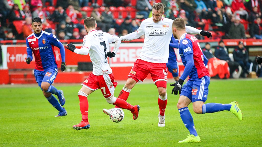 Es gibt ein Fußballspiel. Die Spieler tragen rote und blaue Trikots. Die Fans schauen im Hintergrund zu.