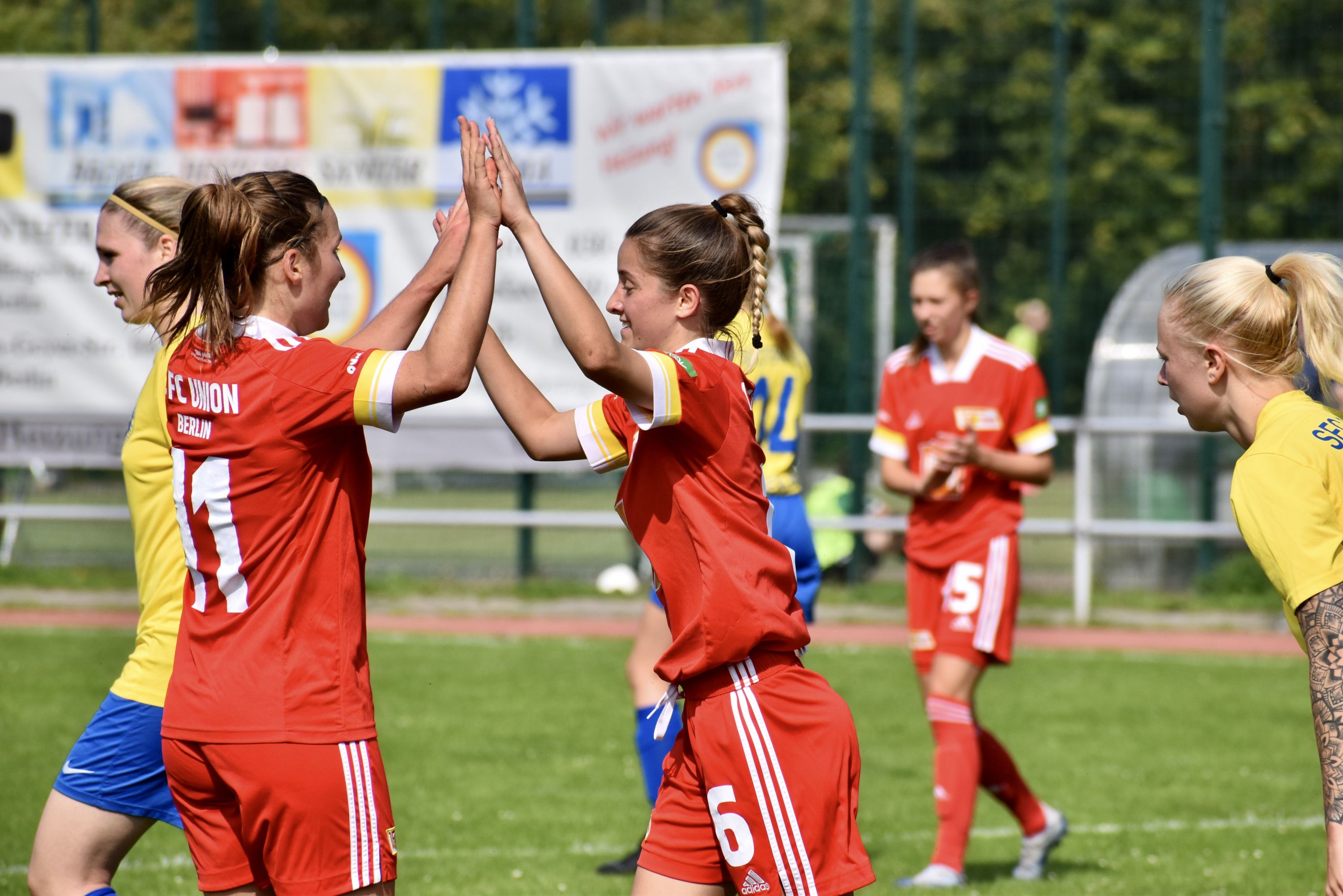 Zwei Fußballspielerinnen in roten Trikots feiern einen gelungenen Spielmoment mit High-Fives auf dem Spielfeld.