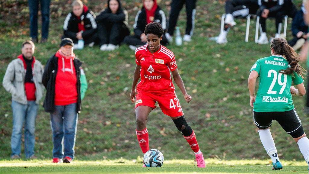Eine Fußballspielerin in rotem Trikot dribbelt den Ball auf einem Spielfeld, während Zuschauer im Hintergrund zuschauen.