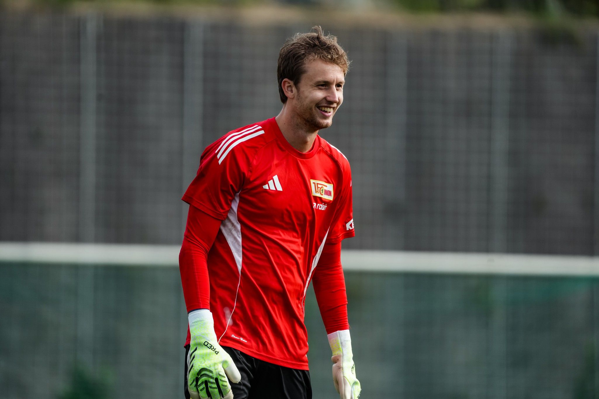 Lächelnder Torwart in rotem Trikot mit weißen Handschuhen, steht auf einem Fußballplatz.