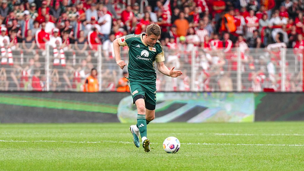 A soccer player in green clothing is kicking the ball on a field, with a cheering crowd in the background.