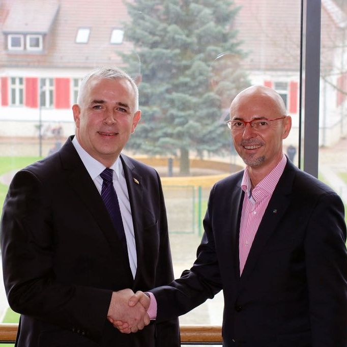 Zwei Männer in Anzügen schütteln sich die Hände vor einem großen Fenster mit Sicht auf einen Garten.