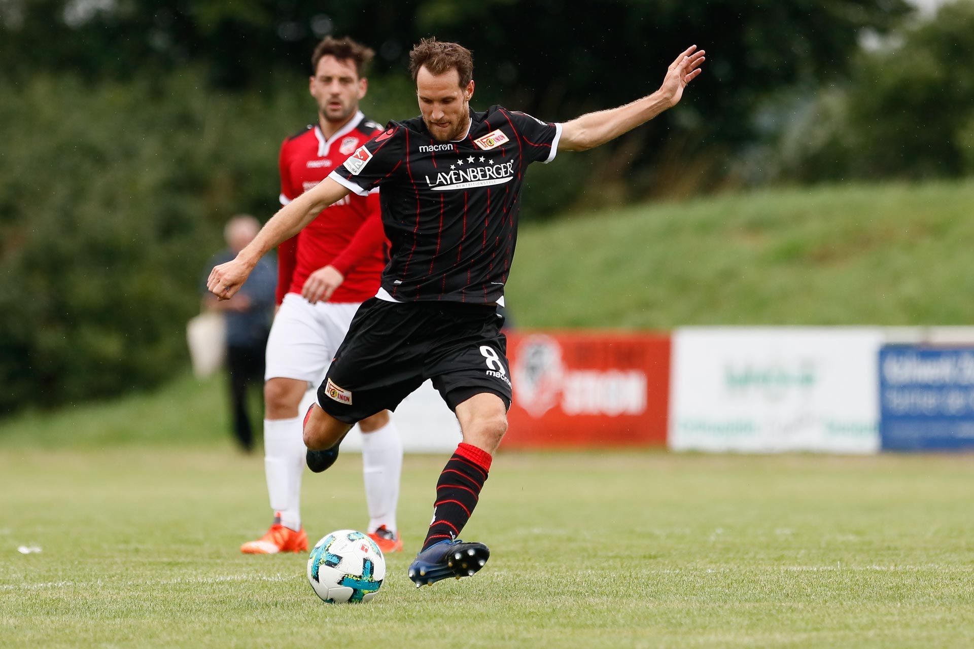 Ein Spieler in einem schwarz-roten Fußballtrikot tritt den Ball auf einem grünen Rasenfeld. Im Hintergrund ist ein anderer Spieler zu sehen.