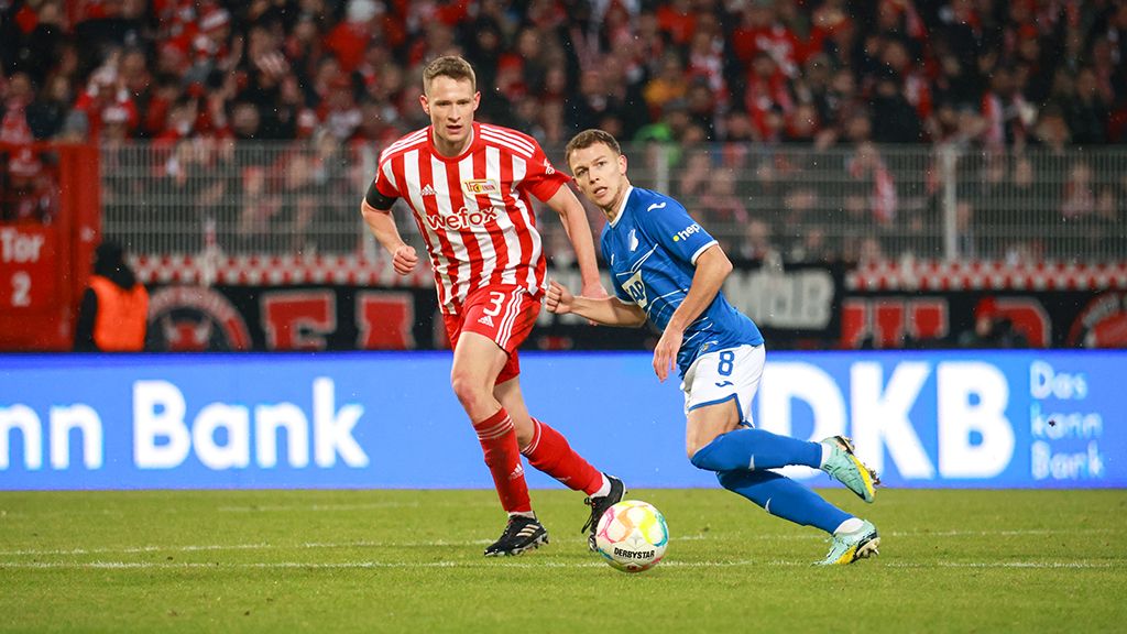 Two soccer players face each other on the field with a ball between them. Spectators are visible in the background.