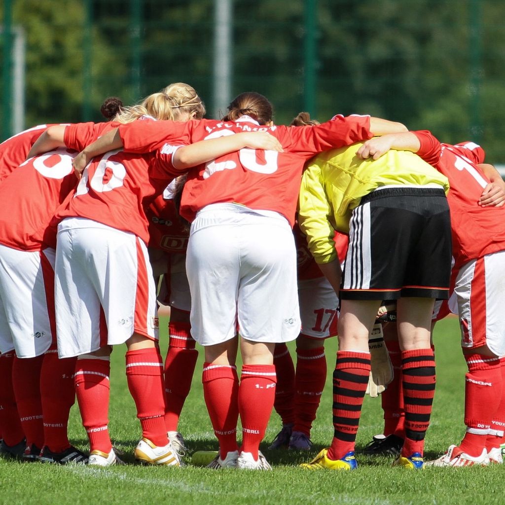 Eine Gruppe von Fußballspielerinnen in roten Trikots bildet einen Huddle auf einem grünen Sportfeld.
