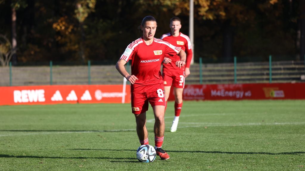 Ein Fußballspieler in rotem Trikot dribbelt den Ball auf einem Trainingsplatz, während ein weiterer Spieler im Hintergrund steht.