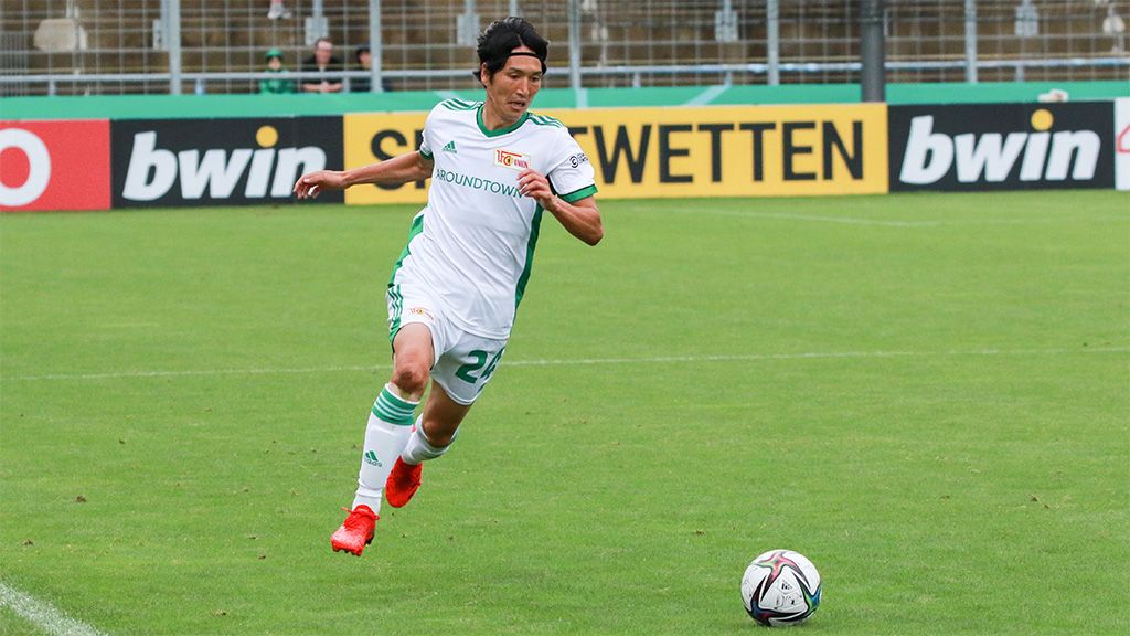 Spieler in weißem Fußballtrikot mit grünen Akzenten dribbelt den Ball auf einem Rasenfeld während eines Spiels.