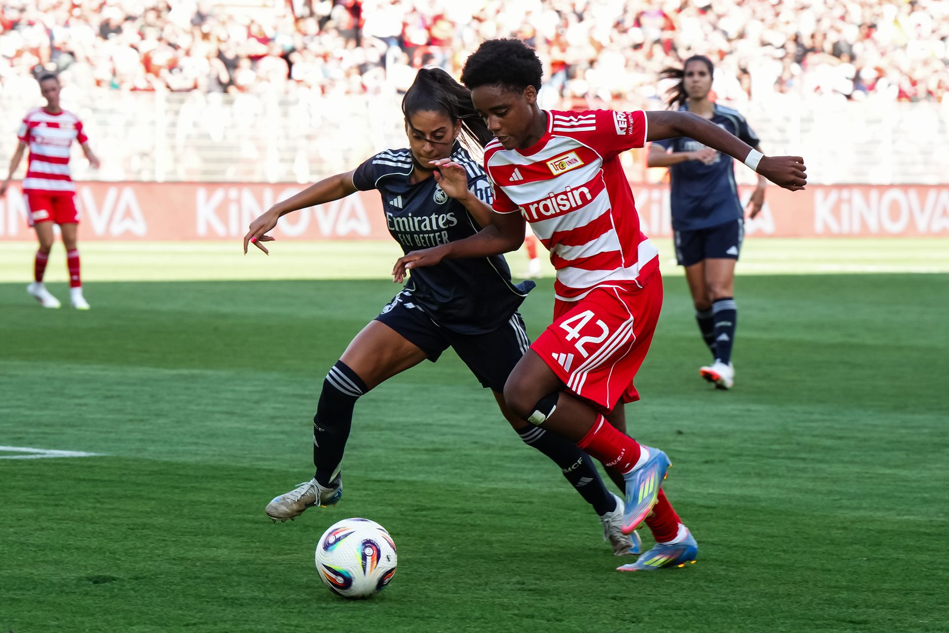Zwei Fußballspielerinnen kämpfen um den Ball auf dem Spielfeld während eines Spiels, umgeben von Zuschauern.