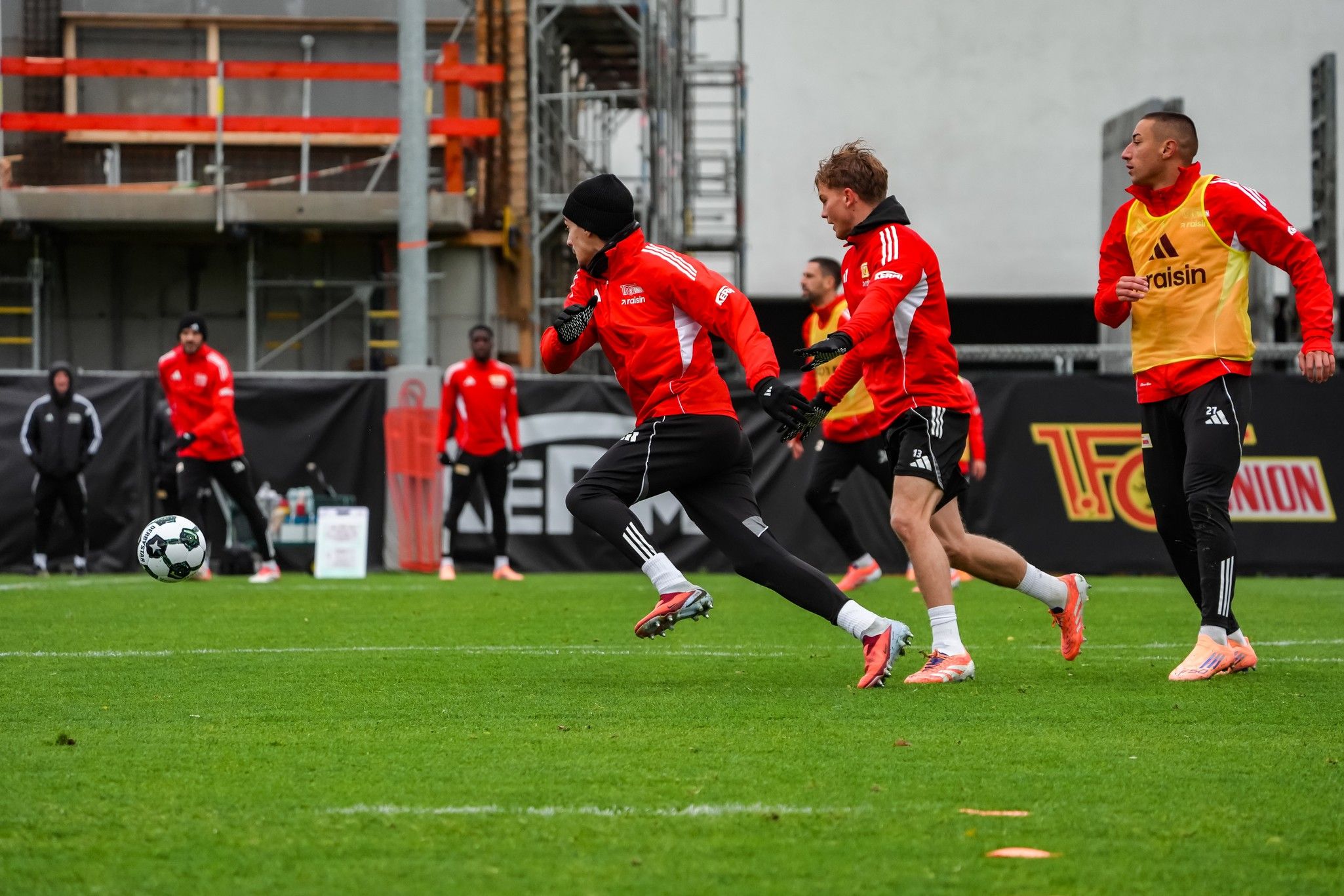 Fußballspieler in roter Kleidung trainieren auf einem grünen Platz, während Zuschauer im Hintergrund stehen.