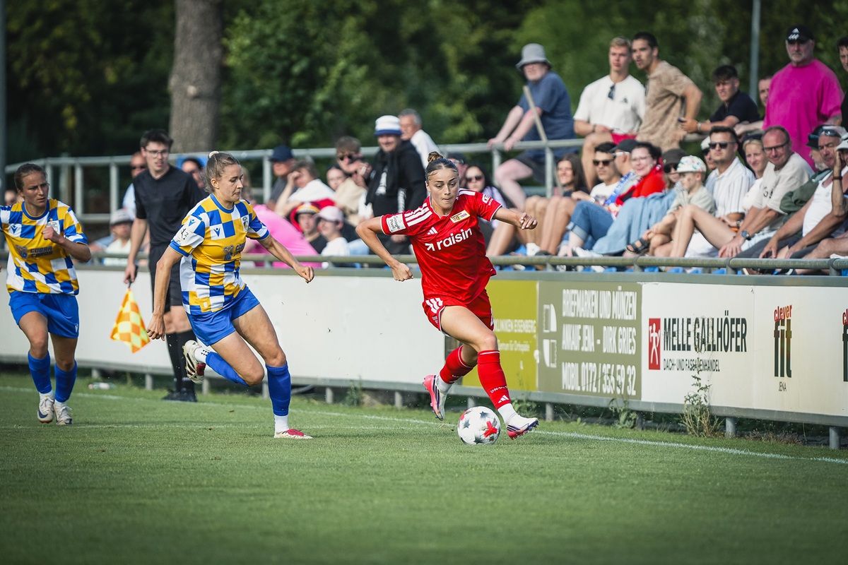 Eine Fußballspielerin in roter Kleidung sprintet mit dem Ball über das Feld, während zwei Spielerinnen in blauen und gelben Trikots sie verfolgen.