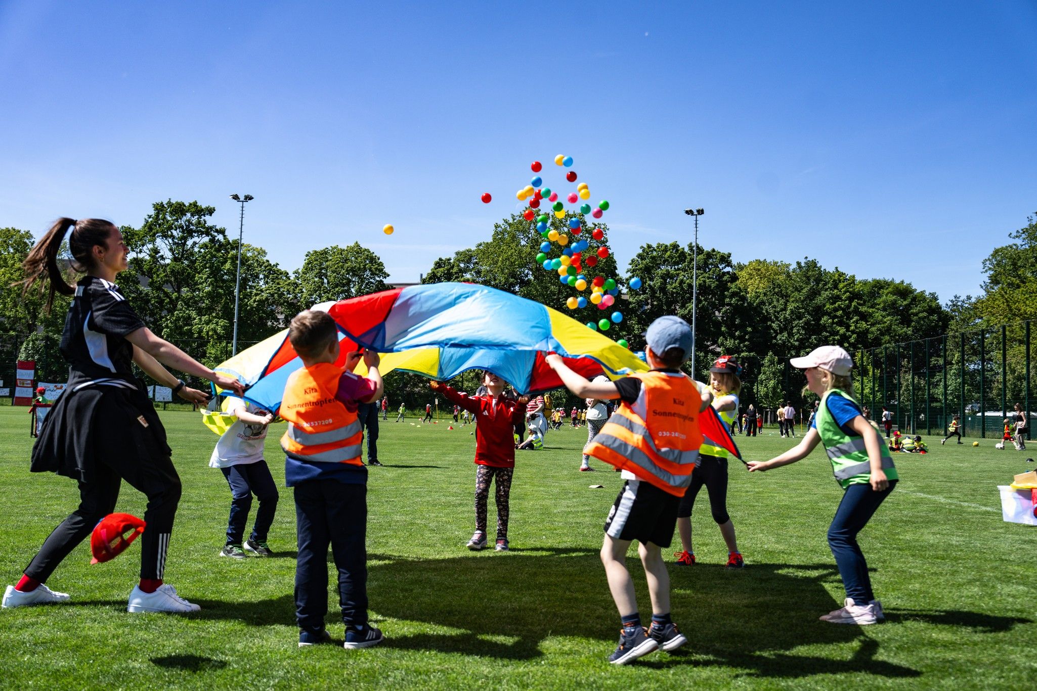 Kinder in bunten Westen spielen mit einem großen, farbigen Fallschirm auf einem grünen Sportfeld, während Luftballons darüber fliegen.