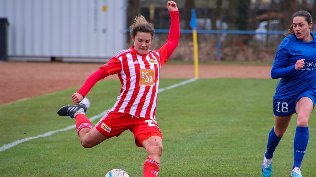 Eine Fußballspielerin in roter und weißer Uniform schießt den Ball, während eine Gegnerin in blauer Uniform in der Nähe steht.