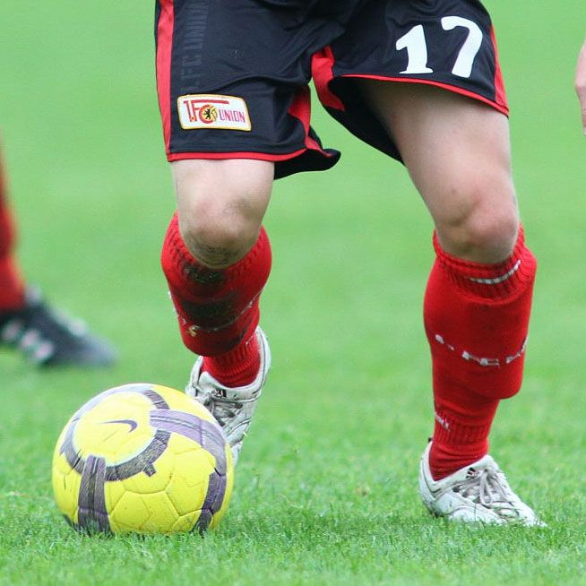 Fußballspieler in roten Shorts und Socken, der gerade einen gelben Fußball auf dem Rasen kickt.