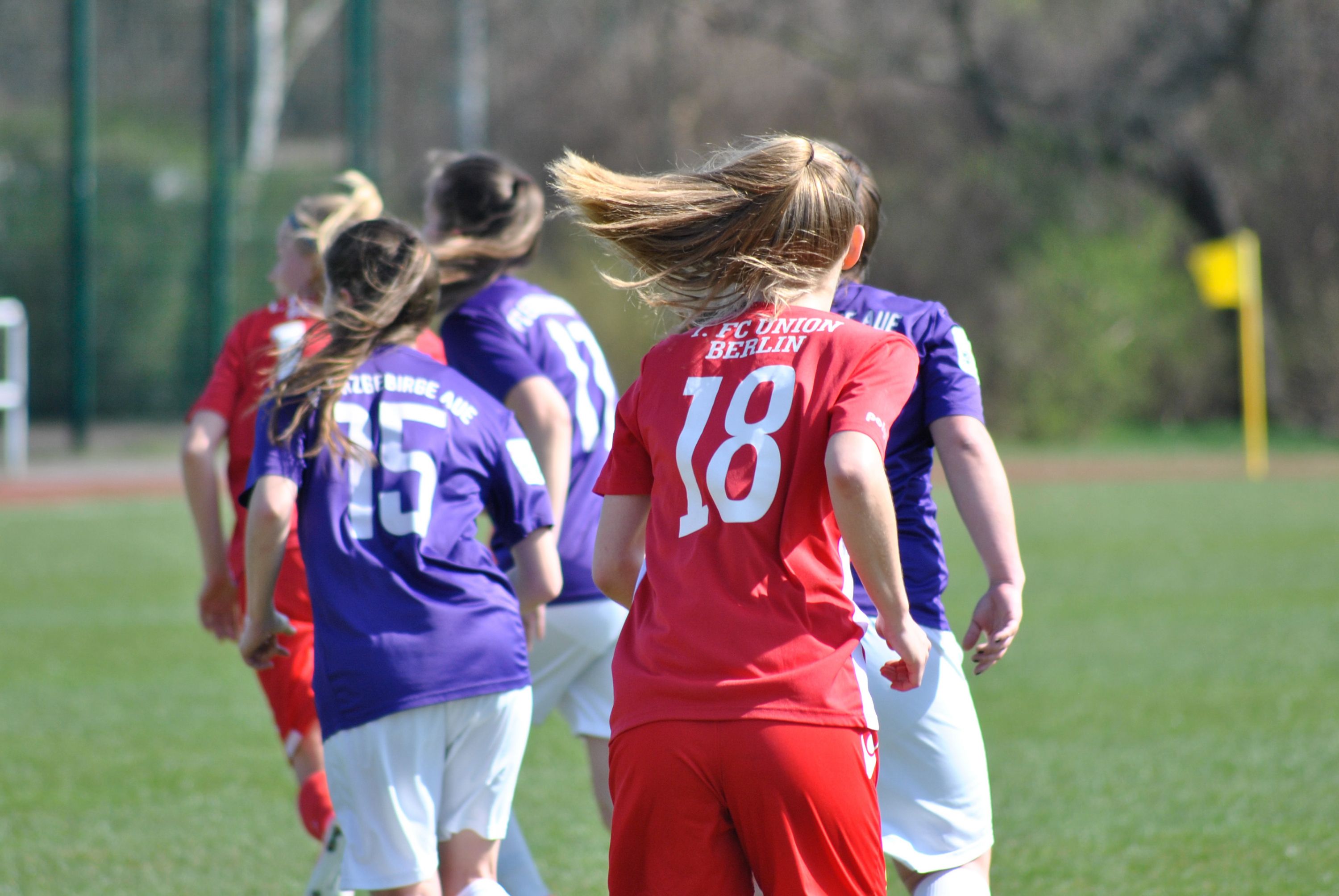 Zwei Gruppen von Fußballspielern in bunten Trikots laufen auf einem grünen Feld in Richtung einer Spielaktion.